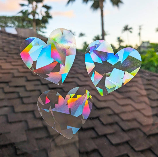 prismatic heart-shaped set of 3 rainbow making suncatcher stickers on a window looking out onto a rooftop and palm trees in Hawaii